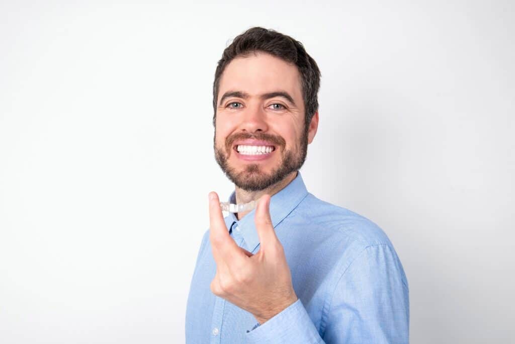 Businessman wearing blue t-shirt over white background holding an invisible aligner ready to use it. Dental healthcare and confidence concept
