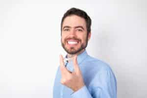 Businessman wearing blue t-shirt over white background holding an invisible aligner ready to use it. Dental healthcare and confidence concept
